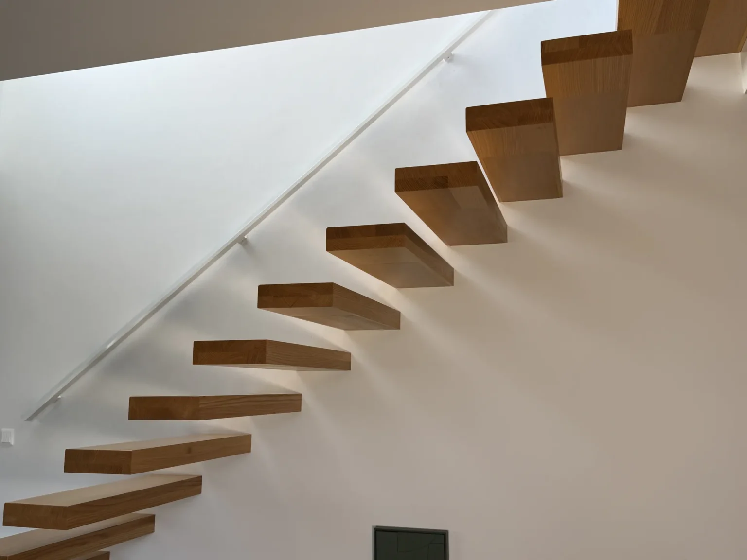 Eine minimalistische, an einer weißen Wand montierte schwebende Holztreppe mit einem eleganten Metallhandlauf, die ein modernes und luftiges Raumdesign schafft.