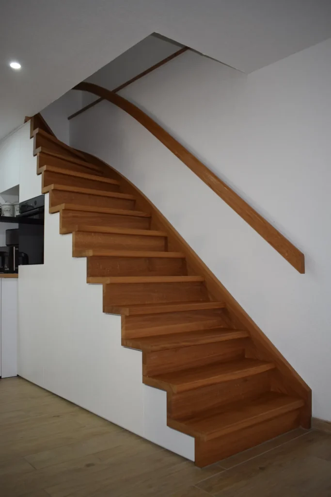 Eine moderne Innentreppe mit Holzstufen und einem geschwungenen Holzhandlauf vor einer weißen Wand, unter der Treppe ist teilweise ein Küchenbereich zu sehen.