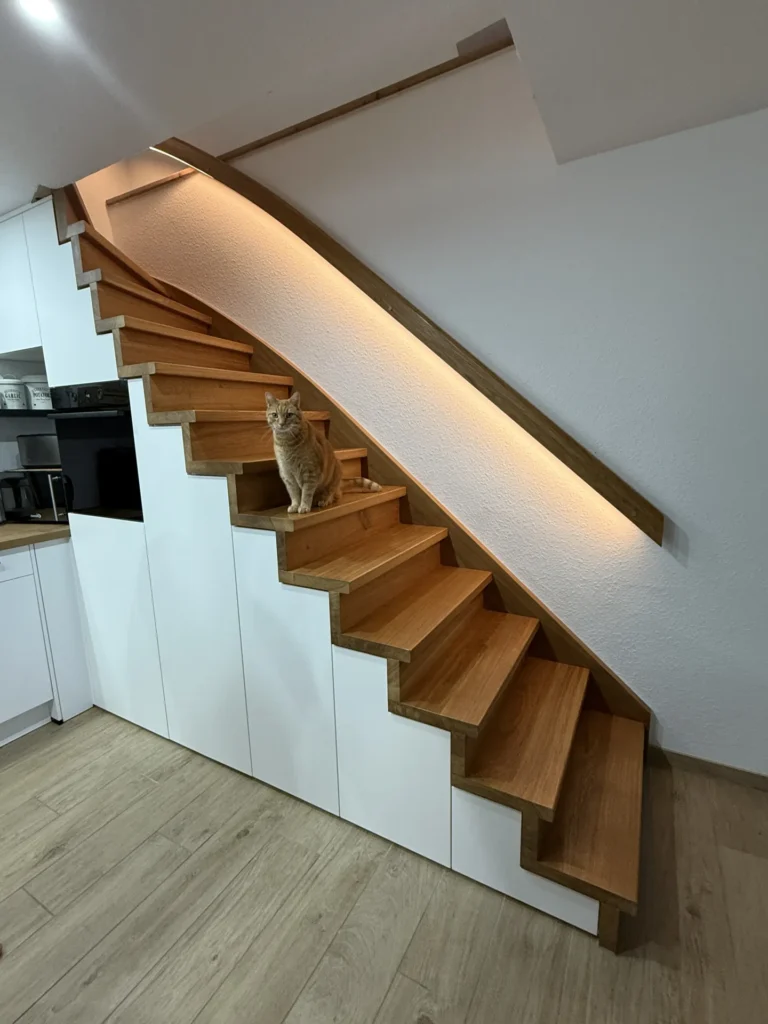 Eine rotbraune Katze sitzt auf halber Höhe einer modernen Holztreppe mit integrierter Beleuchtung. Unter der Treppe stehen weiße Schränke, und der Boden besteht aus hellem Holz.