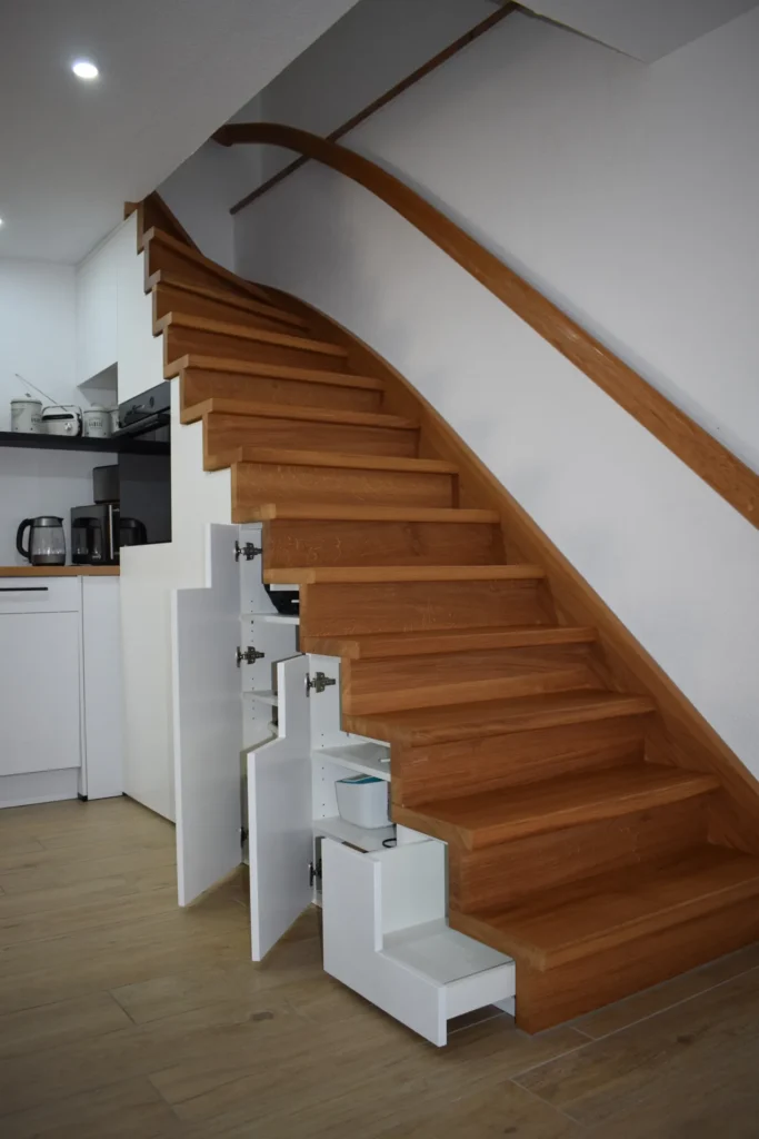 Holztreppe mit eingebauten weißen Schubladen unter den Stufen, die sich neben einem modernen Küchenbereich befindet, in dem auf der Arbeitsplatte Geräte wie eine Kaffeemaschine und eine Mikrowelle stehen.
