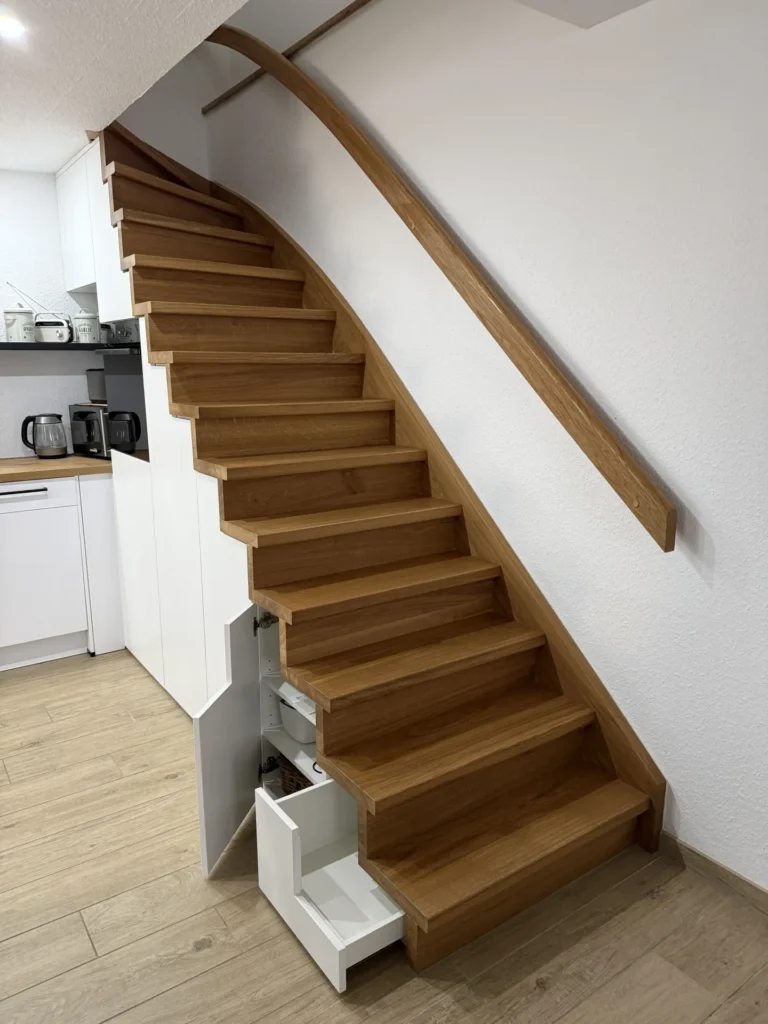 Eine Holztreppe mit einem glatten Handlauf, die über weißen Küchenschränken angebracht ist. Einige Schubladen unter der Treppe sind herausgezogen und geben den Blick auf versteckten Stauraum frei.