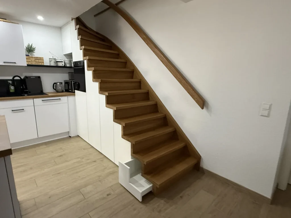 Eine Holztreppe in einer modernen Küche verfügt unten über integrierte weiße Stufen, die als Platz für Haustiere oder als Stauraum dienen. Die Küche ist mit weißen Schränken und einem hellen Holzboden ausgestattet.