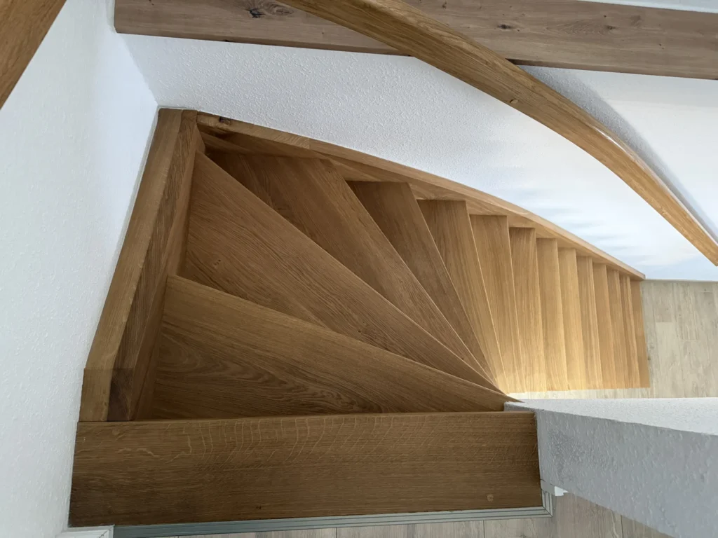 Blick von oben auf eine hölzerne Wendeltreppe mit dreieckigen Stufen, weißen Wänden und einem geschwungenen Holzhandlauf auf der rechten Seite. Die Treppe führt hinunter zu einem hellen Holzboden.