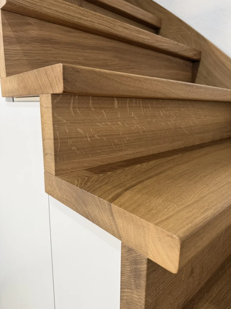 Nahaufnahme einer modernen Holztreppe mit klaren Linien, die die detaillierte Holzmaserung und die scharfen Winkel zeigt, vor einer weißen Wand und einer weißen Tür im Hintergrund.