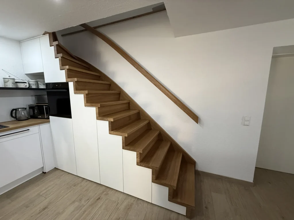 Eine Holztreppe mit weißen Stauschränken darunter in einer modernen Küche mit hellem Holzboden. Die Treppe verfügt über einen schlichten Handlauf und führt an einer weißen Wand entlang nach oben.