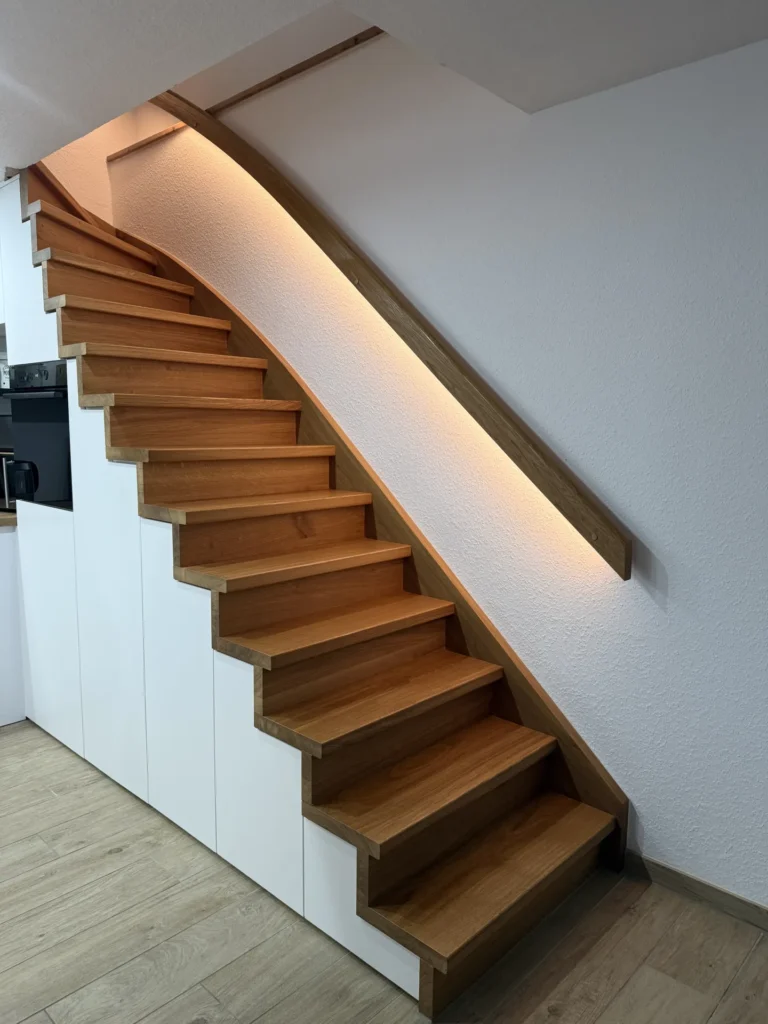 Eine Holztreppe mit integriertem Stauraum darunter, ausgestattet mit einem eleganten Handlauf und sanfter LED-Beleuchtung entlang der Wand, in einem modernen Interieur mit weißen Wänden und Holzboden.