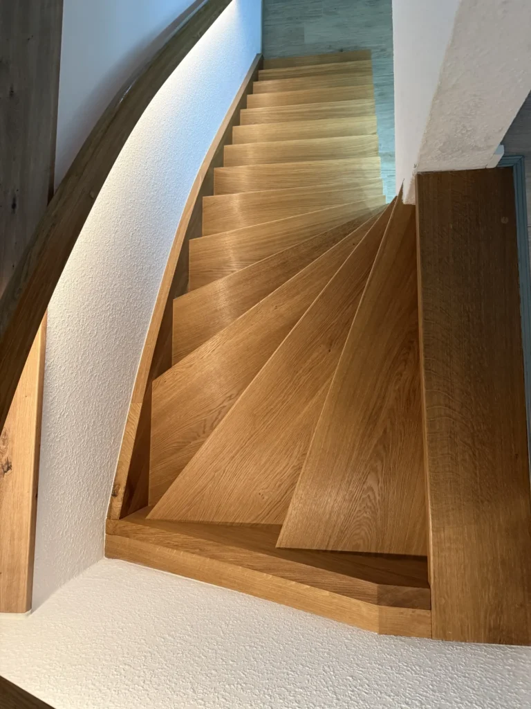 Eine Ansicht von oben auf eine Holztreppe mit schrägen Stufen, die von weißen, strukturierten Wänden und einem Holzhandlauf eingerahmt wird und zu einem hellen Boden hinabführt.