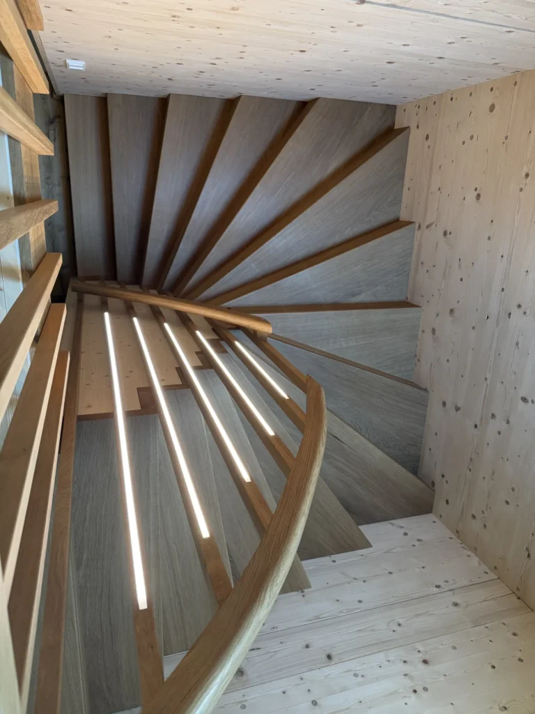 Eine spiralförmige Holztreppe, durch deren Geländer vertikale Lichtstrahlen scheinen; umgeben von hellen Holzwänden und Stufen, die eine moderne und warme Atmosphäre schaffen.