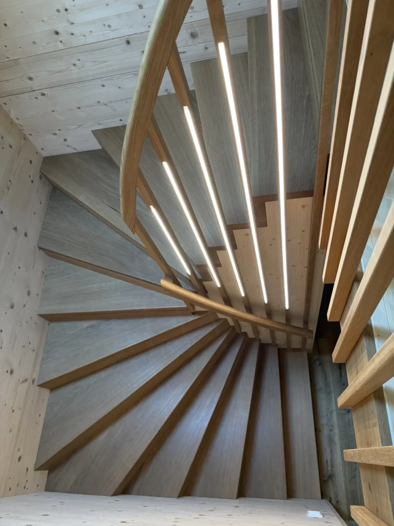 Spiraltreppe aus Holz mit vertikalen LED-Leuchten, die in das Geländer eingelassen sind, von oben gesehen. Helle Holzwände und ein klares, modernes Design sorgen für einen hellen, zeitgemäßen Look.