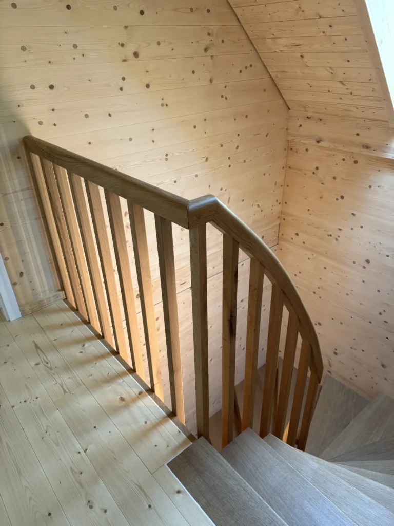 Holztreppe mit passendem Holzhandlauf und vertikalen Balustern, umgeben von hellen Holzwänden und -böden. Natürliches Licht fällt durch ein Fenster darüber herein und erhellt das Treppenhaus.