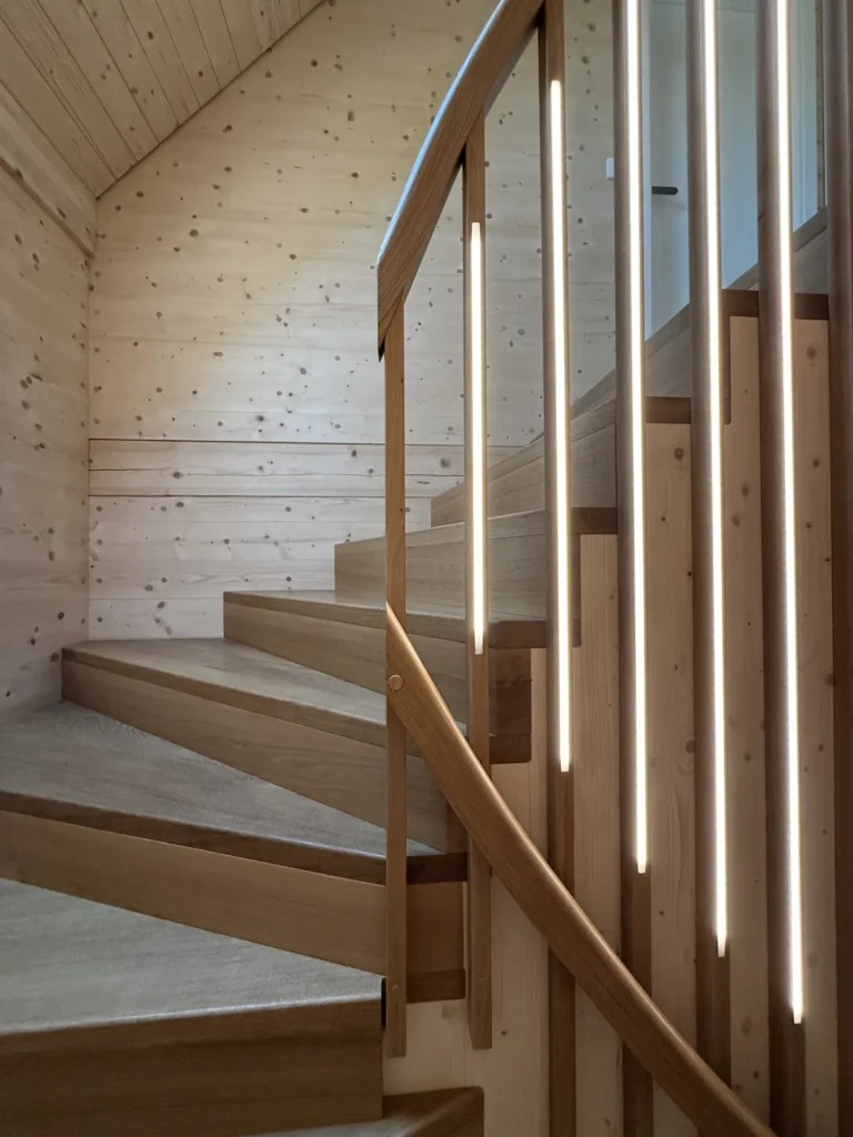 Eine moderne Holztreppe mit vertikal in das Geländer integrierten Leuchten, die vor hellen Holzwänden und einer hellen Holzdecke eine warme und einladende Atmosphäre schafft.