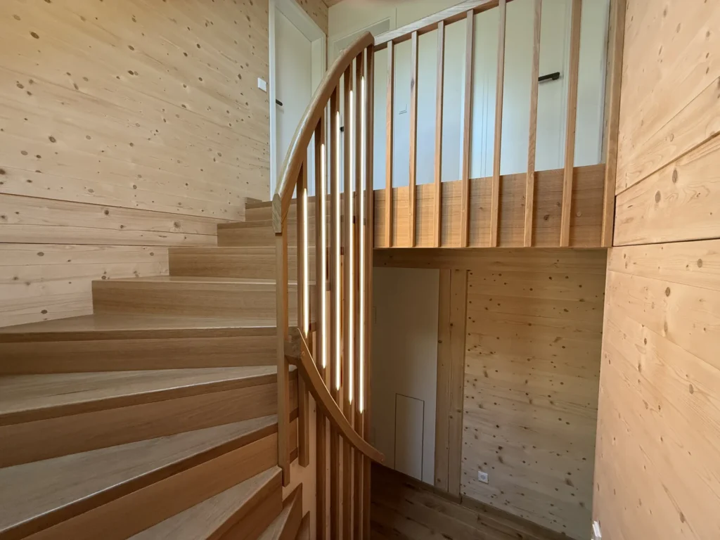 Eine geschwungene Holztreppe mit vertikalen Geländern in einem hellen Raum, dessen Wände, Decke und Boden mit hellem Holz vertäfelt sind. Unterhalb des oberen Treppenabsatzes ist eine Tür zu sehen.