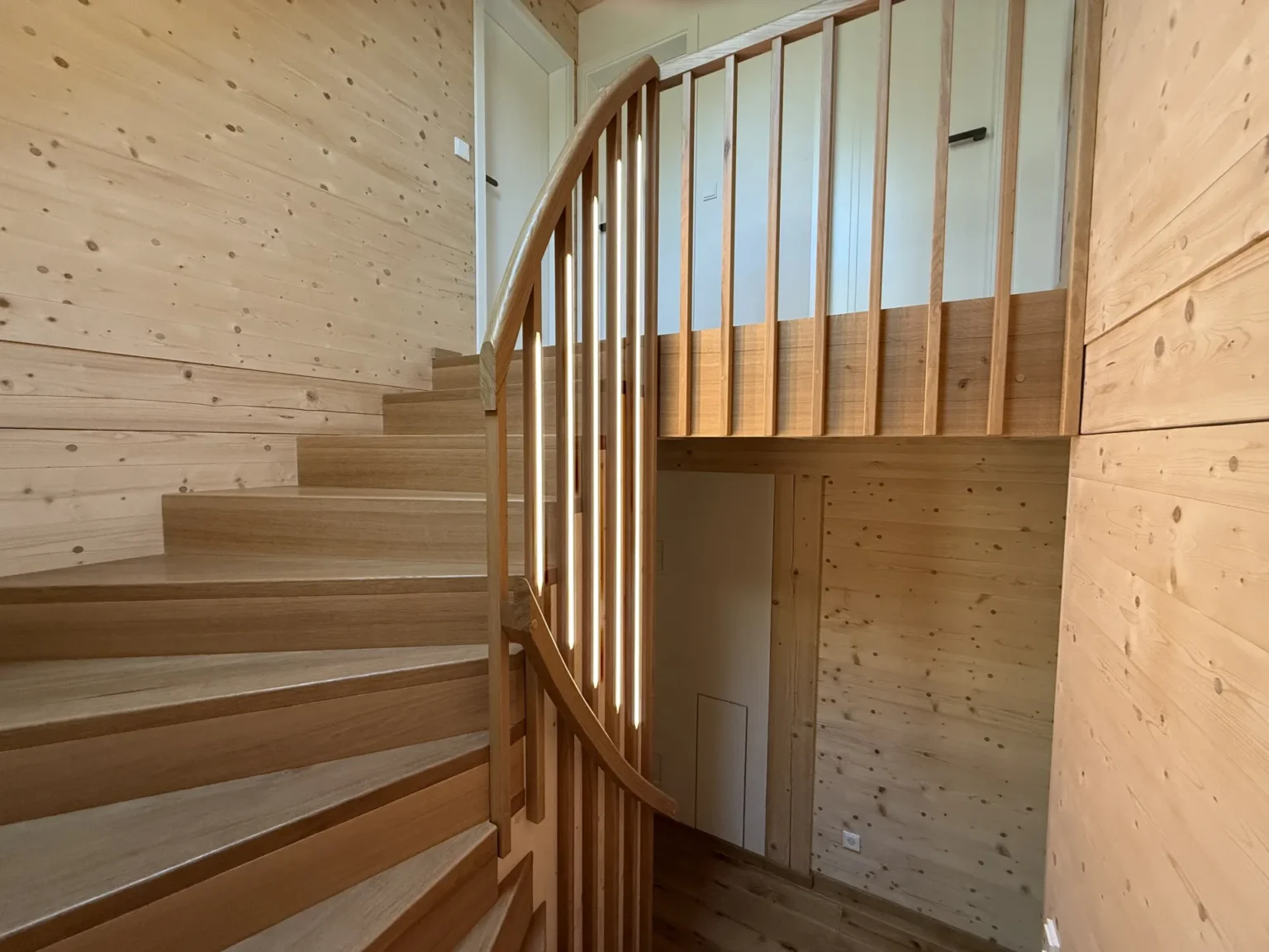 Eine geschwungene Holztreppe mit vertikalen Geländern in einem hellen Raum, dessen Wände, Decke und Boden mit hellem Holz vertäfelt sind. Unterhalb des oberen Treppenabsatzes ist eine Tür zu sehen.