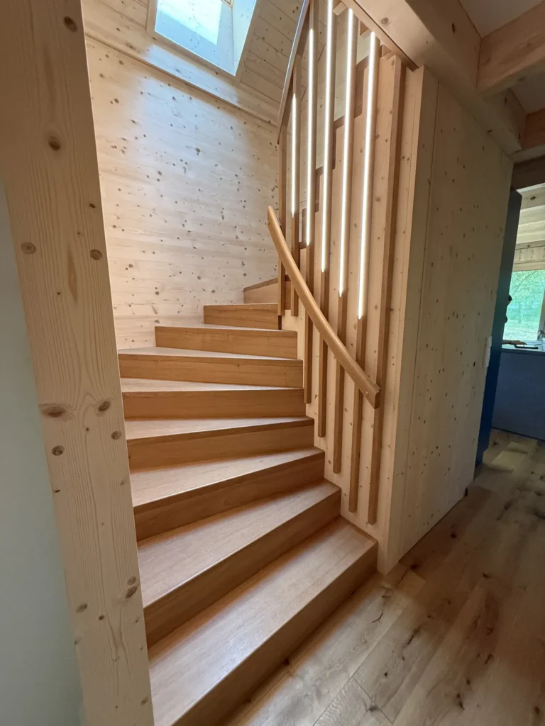 Eine Holztreppe mit geschwungenem Geländer und vertikalen Lamellen führt hinauf zu einem Oberlicht und ist von hellen Holzwänden und -böden umgeben, wodurch im Inneren des Hauses eine warme, natürliche Atmosphäre entsteht.