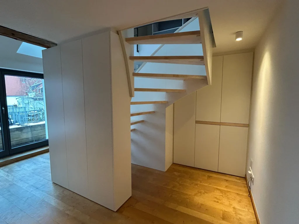 Modernes Interieur mit Holztreppe, weißen Wänden, Einbauschränken und einem großen Fenster auf der linken Seite. Der Raum besticht durch Holzböden und klare, minimalistische Designelemente.