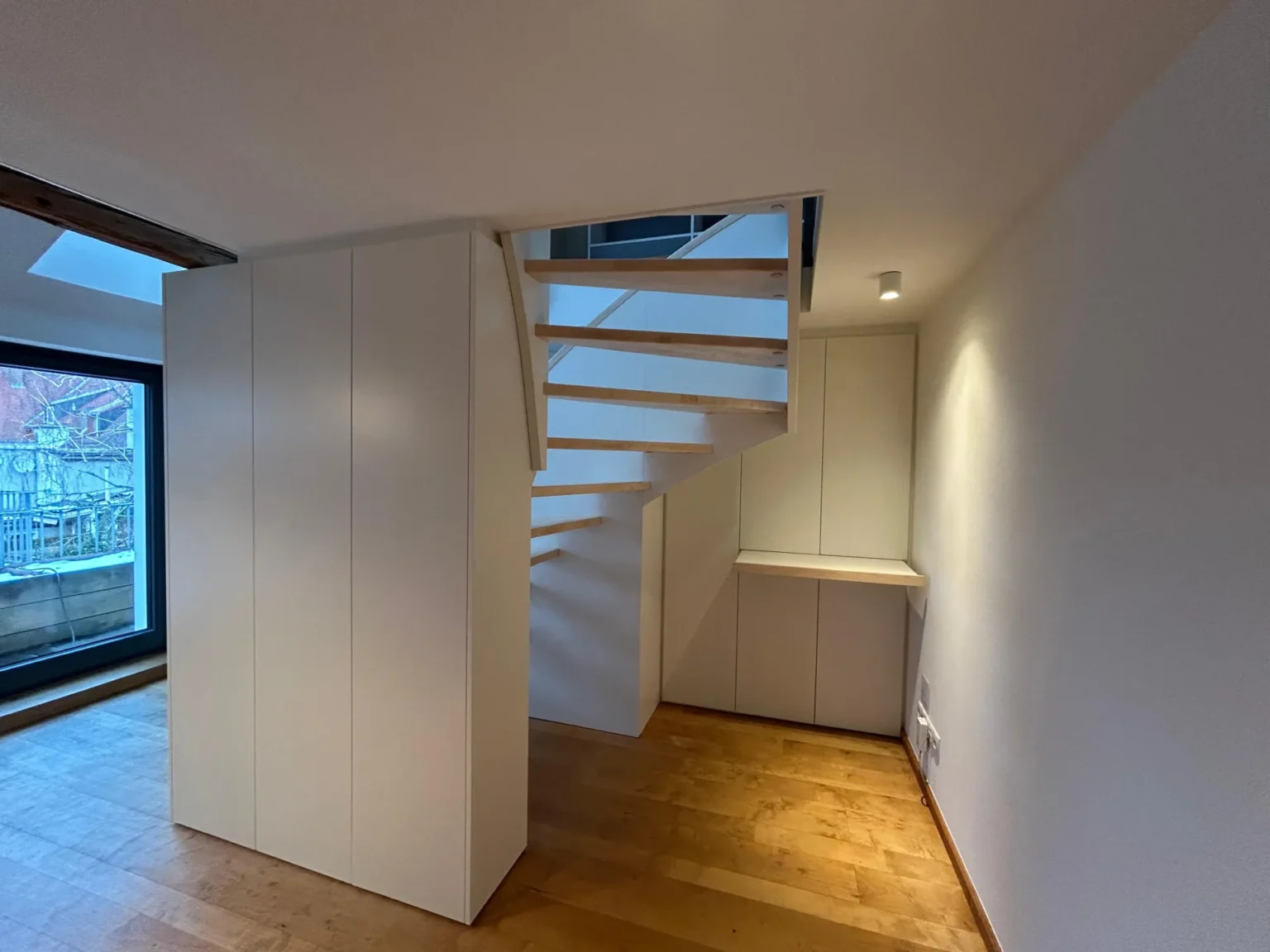 Modernes Interieur mit Holzböden, weißen Wänden und einer offenen Treppe mit Holzstufen. An den Wänden reihen sich Einbauschränke aneinander. Ein großes Fenster auf der linken Seite lässt Tageslicht herein.