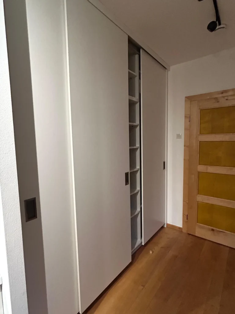 Ein weißer Schrank mit Schiebetür, dessen eine Tür teilweise geöffnet ist und den Blick auf hohe Regale im Inneren freigibt. Der Schrank steht neben einer Holztür mit gelben Füllungen in einem Raum mit Holzboden.