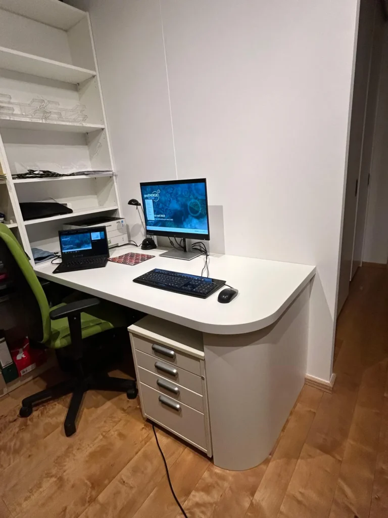 Ein aufgeräumtes Heimbüro mit einem weißen Schreibtisch, einem Computermonitor, einem Laptop, einer Tastatur und einer Maus. Dort stehen ein grüner Stuhl, Regale mit Glaswaren und ein Holzboden. Unter dem Schreibtisch sind Kabel zu sehen.