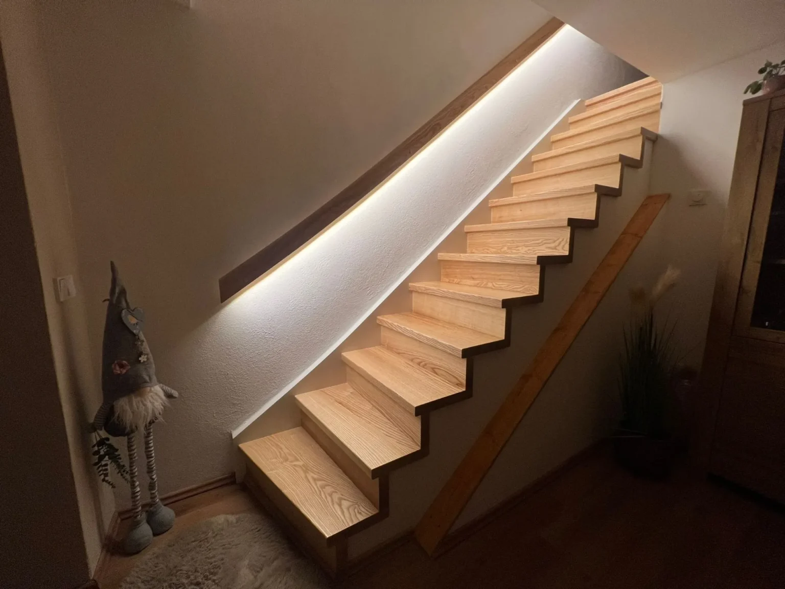 Eine Holztreppe mit LED-Beleuchtung unter dem Handlauf, eine Gartenzwerg-Dekoration, ein kleiner Teppich und eine Topfpflanze am Fuß der Treppe, in einem warm beleuchteten Innenraum.