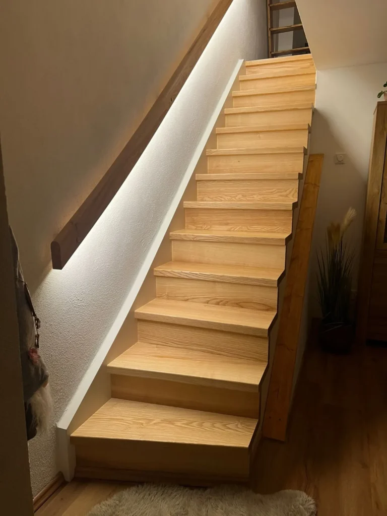 Eine Holztreppe mit integrierter Seitenbeleuchtung an einer weißen Wand, die in einem gemütlichen, modernen Interieur mit Holzböden und Wohnaccessoires in das nächste Stockwerk führt.