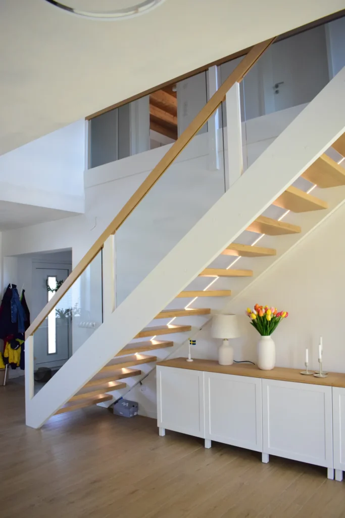 Eine moderne Holztreppe mit Glaseinsätzen, weißen Stufen und integrierter Beleuchtung. Darunter steht ein weißer Schrank, in dem eine Vase mit Tulpen, Kerzen und Dekoartikeln ausgestellt ist. Der Raum ist hell und besticht durch ein minimalistisches, klares Design.