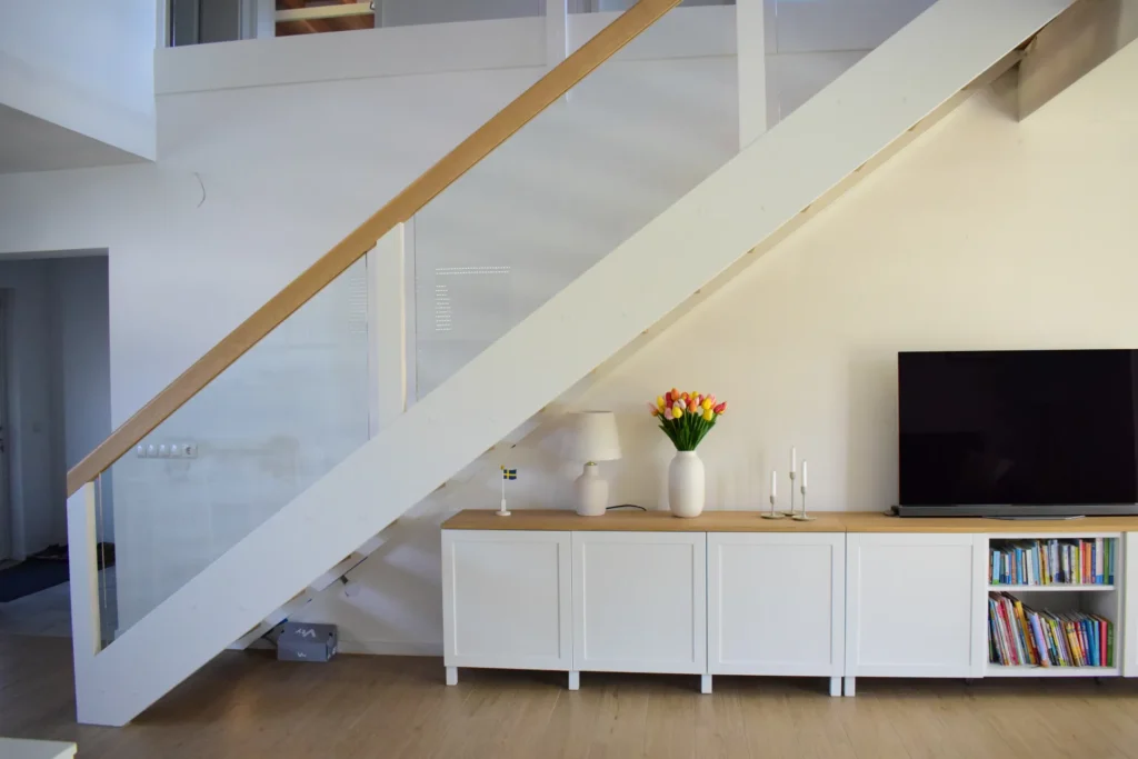 Modernes Wohnzimmer mit einem weißen Schrank unter der Treppe, in dem eine Vase mit Tulpen, Kerzenständer und ein Fernseher stehen. Ein Teil des Schranks ist mit Büchern gefüllt. Helle, minimalistische Einrichtung mit hellen Holzböden.