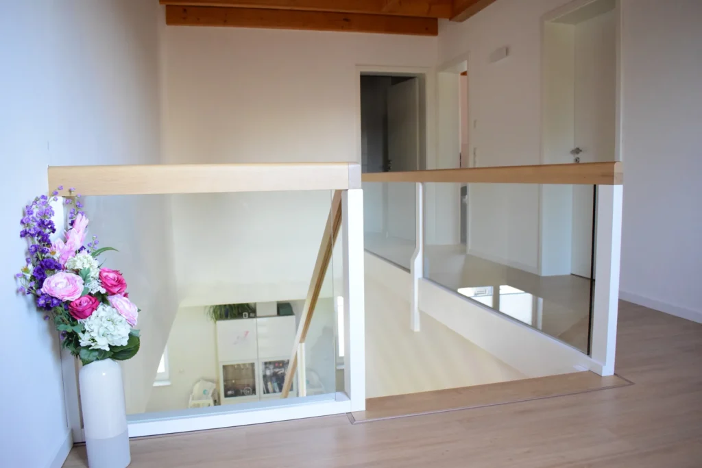 Ein moderner Flur mit hellen Holzböden, weißen Wänden und einem Holzgeländer. Neben einer Glaswand steht neben der Treppe eine weiße Vase mit rosa, violetten und weißen Blumen. Von hier aus führen Türen in andere Räume.