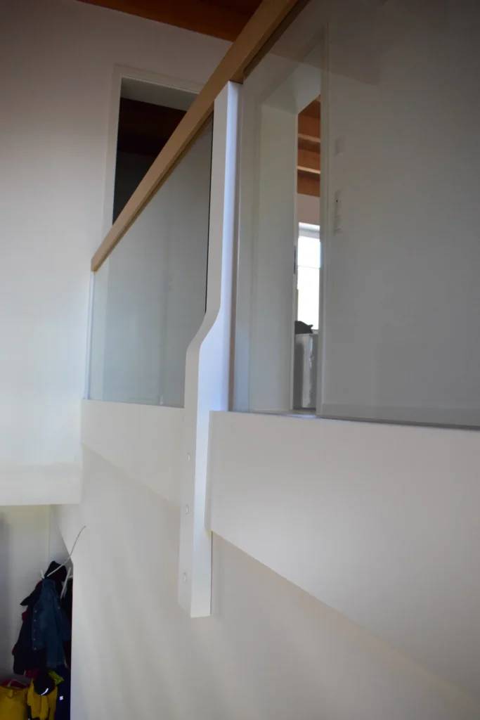 Blick auf einen modernen Innenbalkon mit Glasgeländer und hellen Holzverkleidungen, mit weißen Wänden und einer Türöffnung im Hintergrund. An der Wand auf der unteren Ebene hängen einige Kleidungsstücke oder Gegenstände.