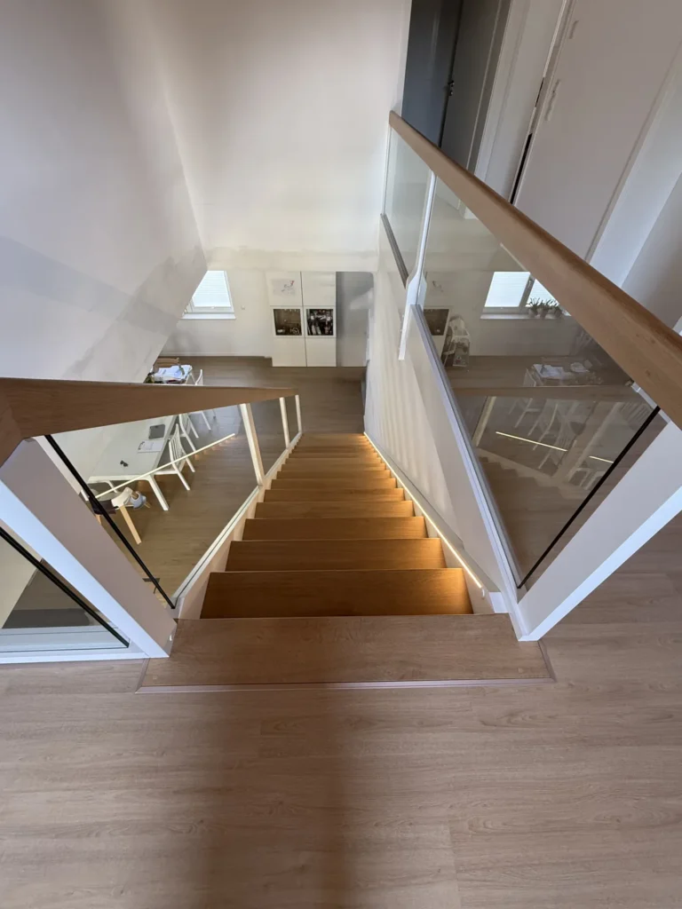 Eine moderne Holztreppe mit Glasgeländern, von oben gesehen. Sanftes Licht beleuchtet die Stufen. Der Boden und die Treppe sind aus hellem Holz, und im Raum darunter sind Möbel zu sehen.