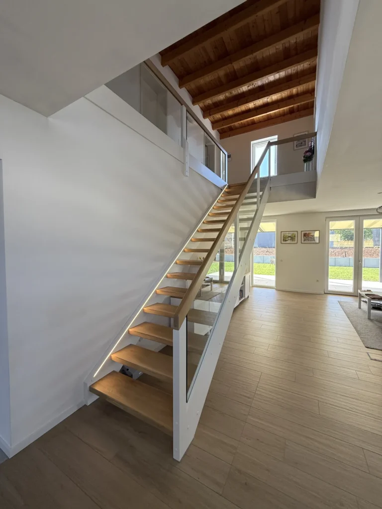 Moderne Innenausstattung mit einer Holztreppe und einem Glasgeländer, die in das Obergeschoss führen; durch die großen Fenster strömt Tageslicht herein, und der Raum ist mit hellem Holzboden und weißen Wänden ausgestattet.