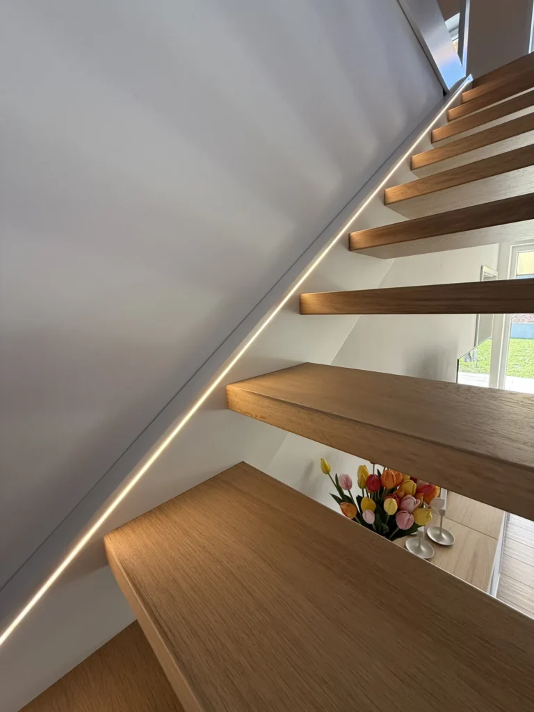 Eine schwebende Holztreppe mit integrierter LED-Beleuchtung entlang der Wand. Unterhalb der Treppe steht eine Vase mit bunten Tulpen. Durch eine Tür im Hintergrund fällt Tageslicht herein und bringt das moderne Interieur zur Geltung.