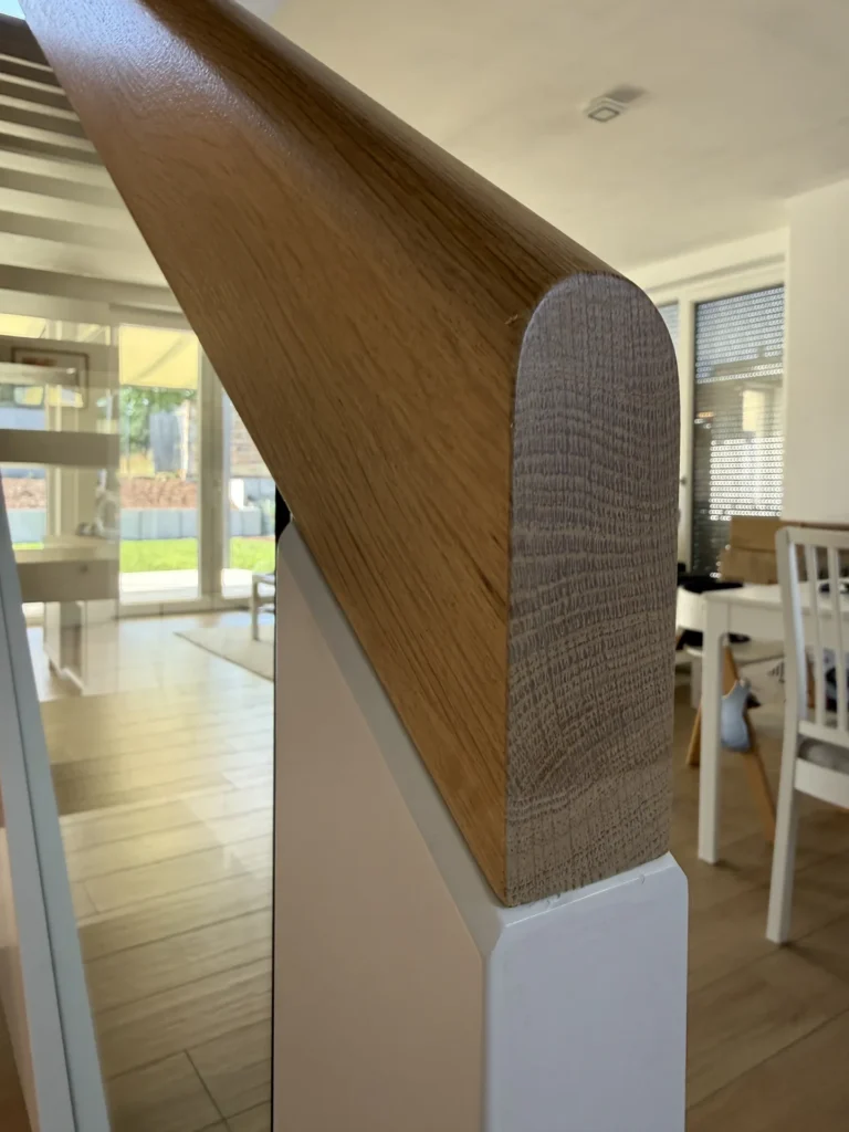 Nahaufnahme eines Holzhandlaufs an einer Treppe in einem modernen, sonnendurchfluteten Innenraum mit weißen Wänden, hellen Holzböden und Möbeln, die im Hintergrund zu sehen sind.