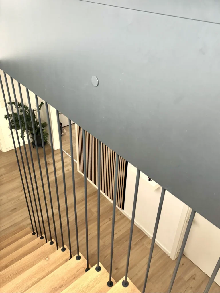 Blick die moderne Treppe hinunter mit Holzstufen und vertikalen schwarzen Metallgeländern. Der Boden besteht aus hellem Holz, und in der Ecke neben einer weißen Wand und einer Tür ist eine grüne Pflanze zu sehen.