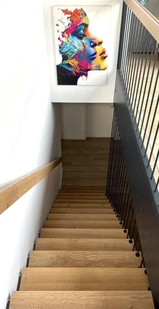 Eine Holztreppe mit schwarzem Metallgeländer führt hinunter zu einem Treppenabsatz. An der weißen Wand am Fuß der Treppe hängt ein großes, farbenfrohes abstraktes Gemälde, das das Gesicht einer Frau darstellt. Von rechts fällt Licht herein.