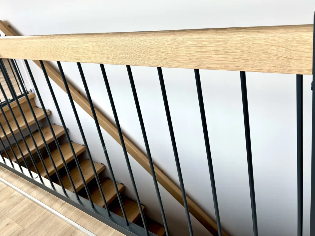 Moderne Treppe mit hellen Holzstufen und schwarzem Metallgeländer, von oben gesehen. Das Geländer besteht aus vertikalen schwarzen Stäben und einem Holzhandlauf und bildet einen Kontrast zur weißen Wand im Hintergrund.