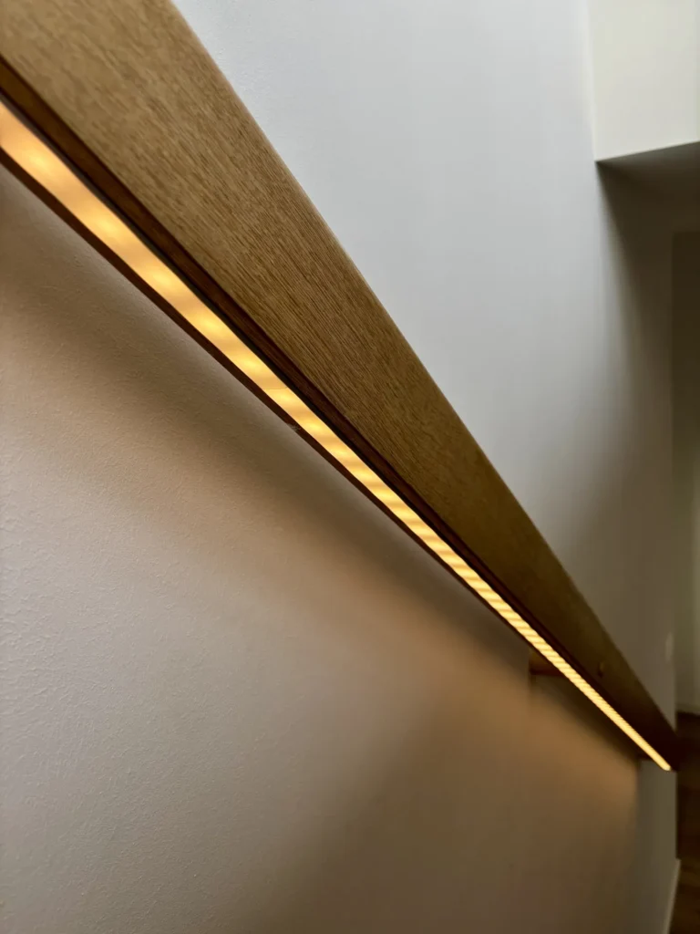 Eine Nahaufnahme eines modernen Holzhandlaufs mit integrierter warmer LED-Beleuchtung, der an einer weißen Wand in einem minimalistisch gestalteten Innenraum angebracht ist.