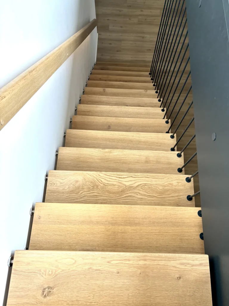 Eine Ansicht von oben auf eine moderne Holztreppe im Innenbereich mit Metallgeländern auf der rechten Seite und einem Holzhandlauf auf der linken Seite, die hinunter zu einem hellen Holzboden führt.