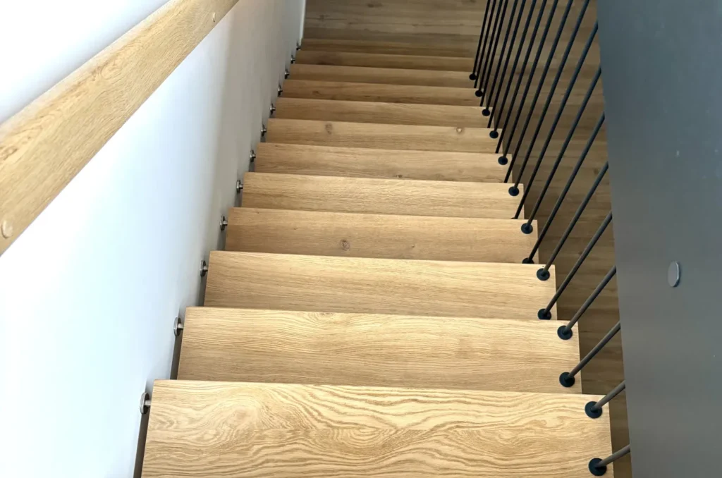 Eine Ansicht von oben auf eine moderne Holztreppe im Innenbereich mit Metallgeländern auf der rechten Seite und einem Holzhandlauf auf der linken Seite, die hinunter zu einem hellen Holzboden führt.