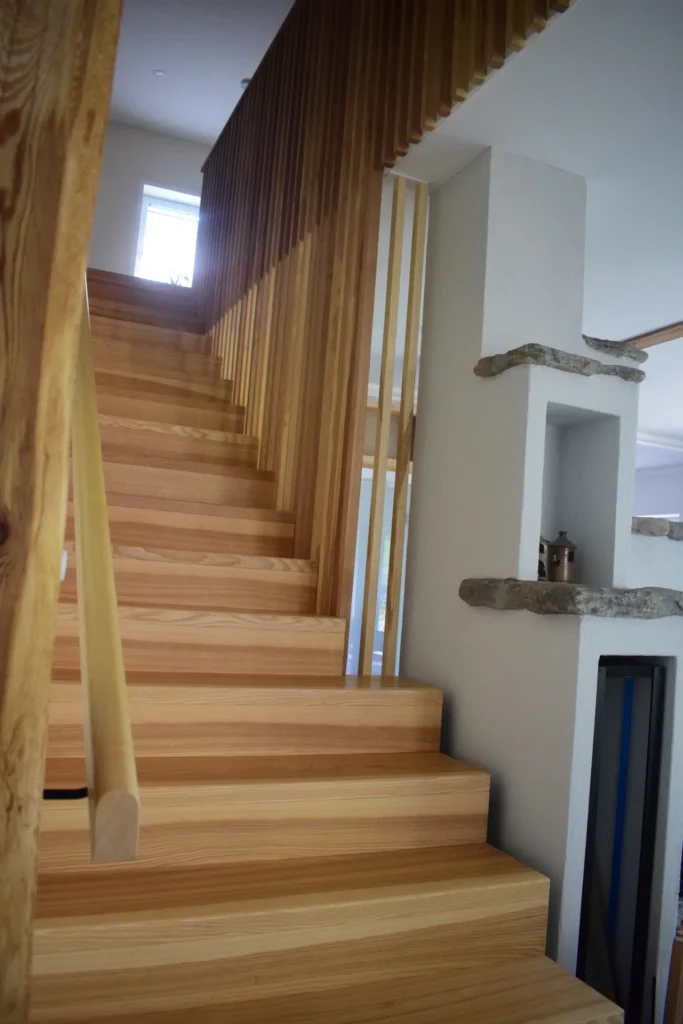 Eine Holztreppe mit vertikalen Lamellen als Geländer, neben einer weißen Wand mit Einbauregalen und Akzenten aus Stein, in einem modernen, hell beleuchteten Innenraum.