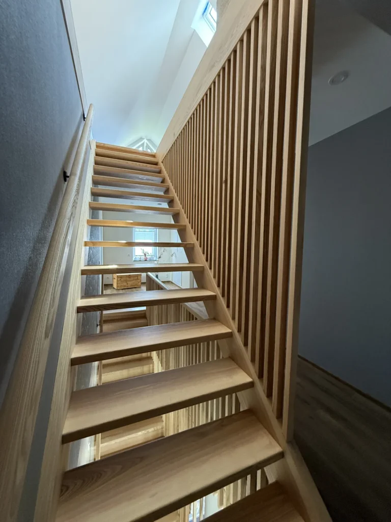 Eine moderne Holztreppe mit offenen Setzstufen und vertikalen Lamellen führt in einem hellen Innenraum nach oben, in den natürliches Licht durch die darüber liegenden Fenster hereinströmt.