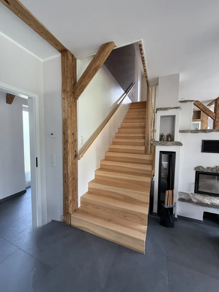 Eine Holztreppe mit Handlauf führt in einem modernen Haus mit sichtbaren Holzbalken, weißen Wänden, dunkelgrauem Fliesenboden und einem Kamin auf der rechten Seite nach oben.