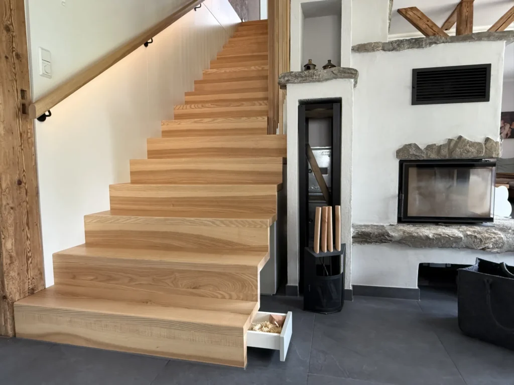 Eine moderne Holztreppe mit einer versteckten Schublade, die sich auf der untersten Stufe öffnet, neben einer weißen Wand, einem Kamin mit Steinakzenten und gestapeltem Brennholz in einem Halter auf einem dunklen Fliesenboden.
