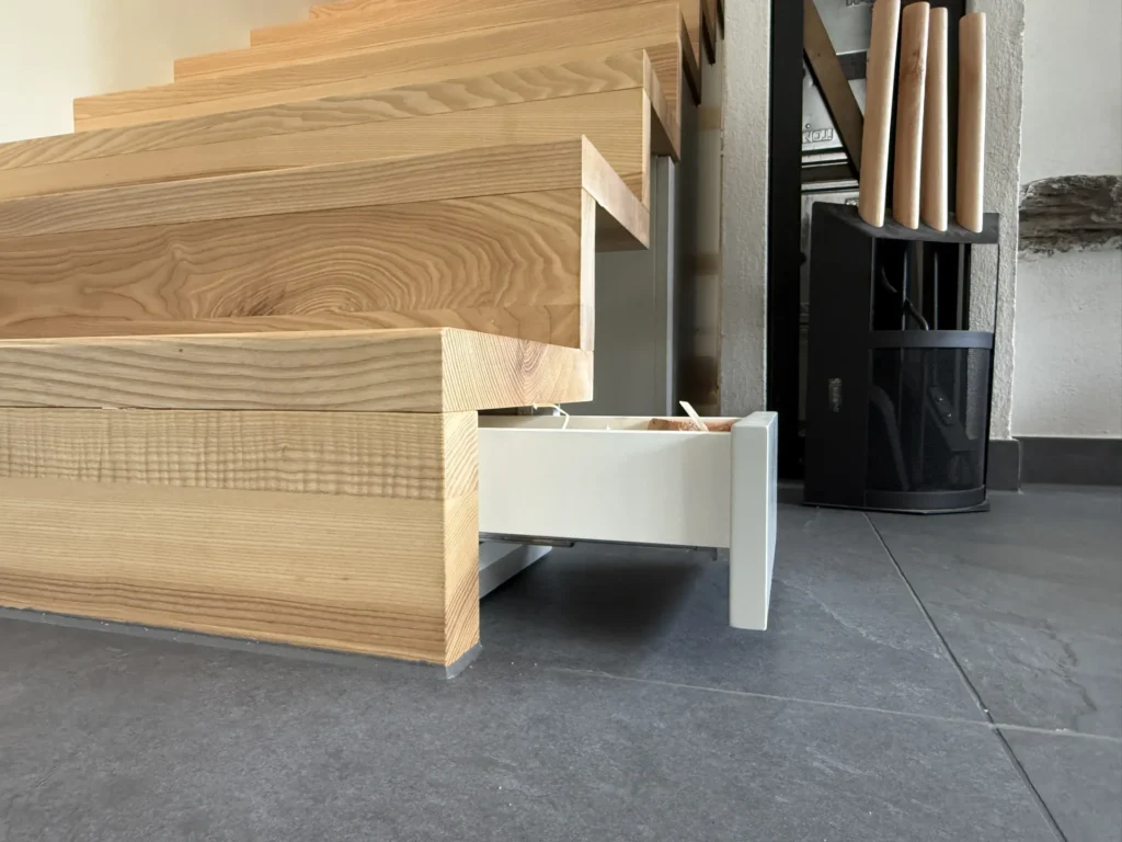 Eine Holztreppe, bei der sich aus der untersten Stufe eine versteckte Schublade herausziehen lässt, die einen raffinierten Stauraum freigibt. Der Boden ist mit dunkelgrauen Fliesen ausgelegt, und in der Nähe stehen Holzgegenstände sowie ein schwarzer Ständer.