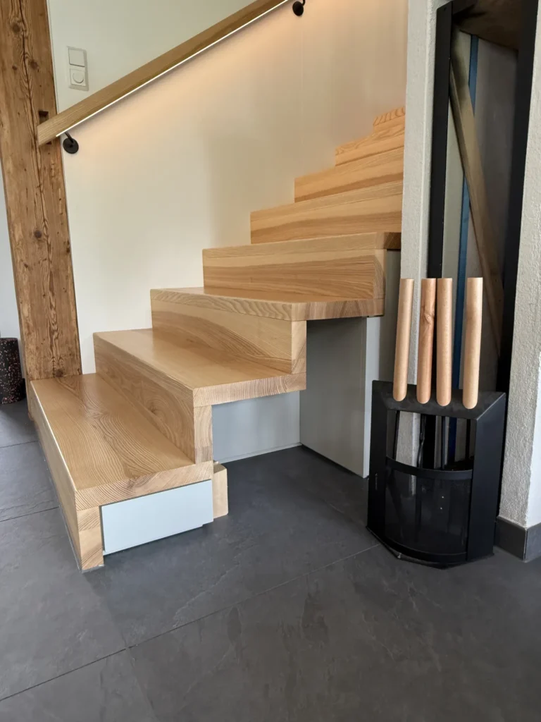 Moderne Holztreppe mit breiten, rechteckigen Stufen und minimalistischem Design. Neben der Treppe steht ein schwarzer Ständer, auf dem vier helle, zylindrische Gegenstände liegen – möglicherweise Kaminbesteck oder Nudelhölzer.