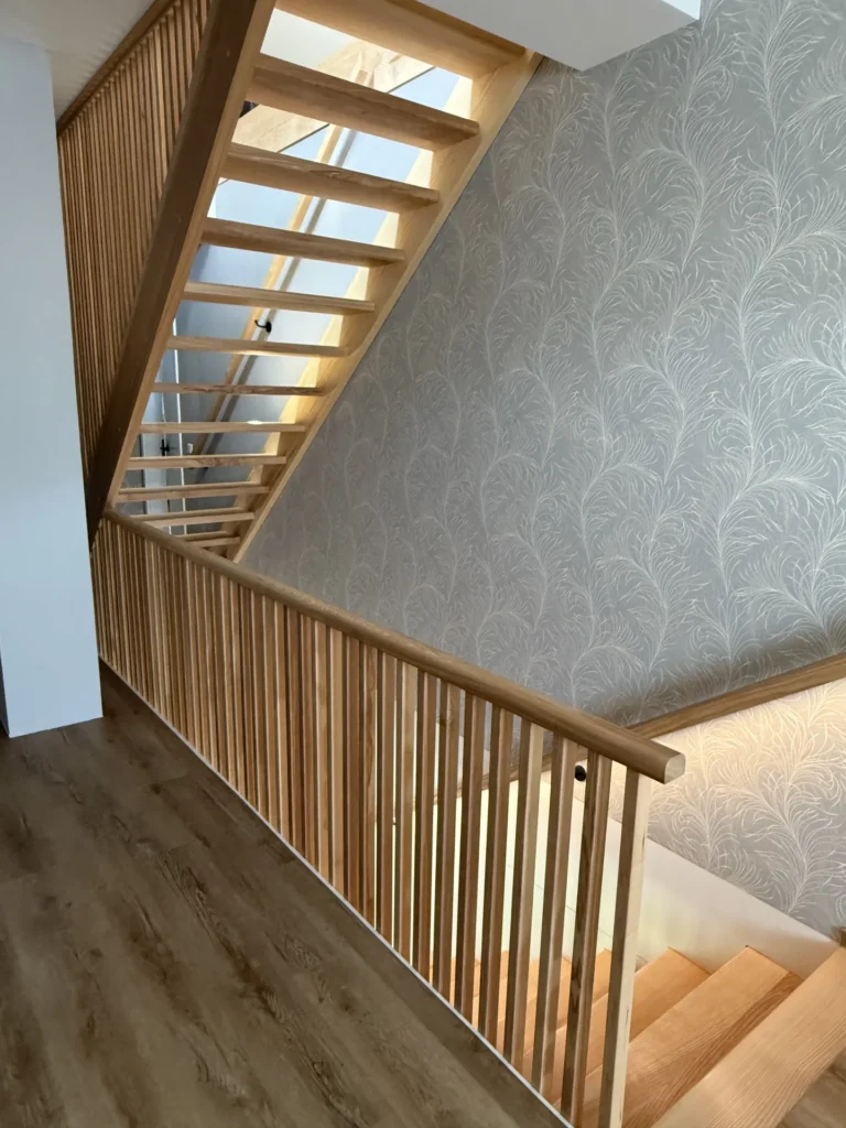 Eine moderne Holztreppe mit vertikalen Lamellen und einer mit gemusterter Tapete versehenen Wand, vom Obergeschoss aus gesehen. Das natürliche Licht bringt die Holzstruktur und die klaren Linien des Raums besonders gut zur Geltung.