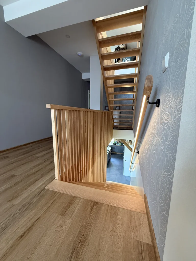 Eine Holztreppe mit vertikalen Lamellen und einem Handlauf führt von einem modernen Innenraum mit hellem Holzboden, grauen Wänden und einer gemusterten Akzentwand hinunter. Eine lineare Wandleuchte beleuchtet das Treppenhaus.