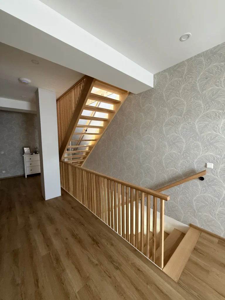 Moderne Innentreppe mit Holzstufen und Geländer, daneben eine hell tapezierte Wand mit Blattmuster sowie ein Holzboden. Am Ende des Flurs ist eine weiße Kommode zu sehen.
