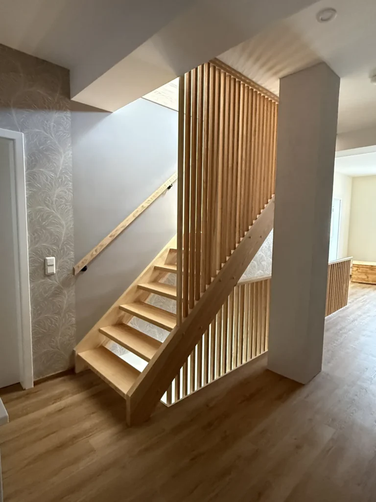 Eine moderne Innentreppe mit Holzstufen und vertikalen Holzlatten als Geländer, eingebettet in einen hellen Raum mit hellen Holzböden und gemusterter Tapete an einer Wand.