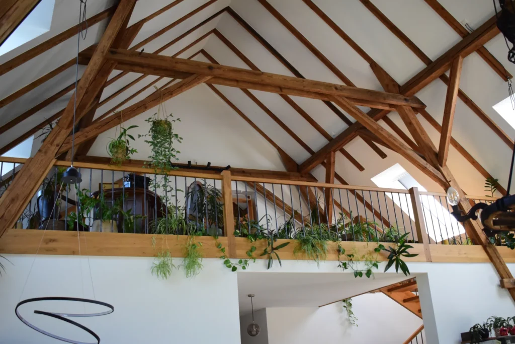Ein Loft mit freiliegenden Holzbalken, Zimmerpflanzen, die an einer Holzgeländerleiste hängen oder darauf stehen, und natürlichem Licht, das durch Oberlichter in der Decke hereinfällt.