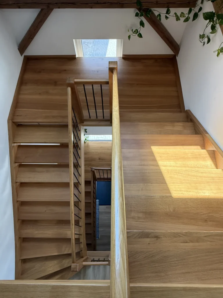 Holztreppe in einem sonnendurchfluteten Innenraum, von oben betrachtet. Die Treppe zeichnet sich durch klare Linien, einen Holzhandlauf, freiliegende Balken, ein kleines Fenster und einige grüne Blattpflanzen aus, die am oberen Rand zu sehen sind.